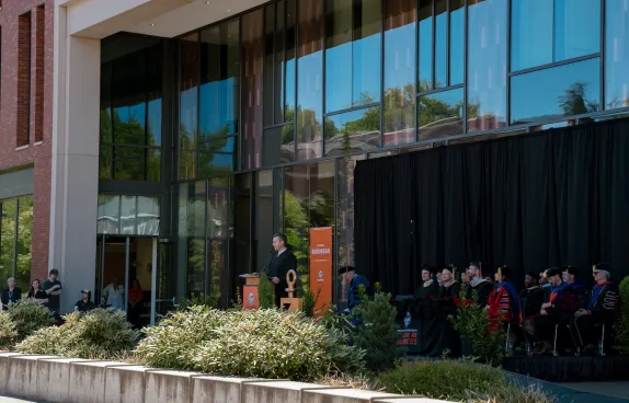 Scott Christianson stands at a podium in front of Austin Hall. He is wearing regalia and delivering a speech. In the background are College of Business faculty and leadership.
