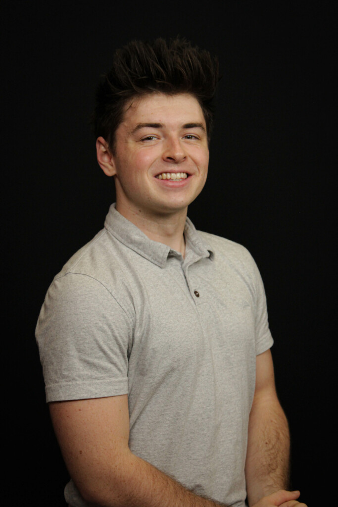 A headshot of a man wearing a gray shirt on a black background.