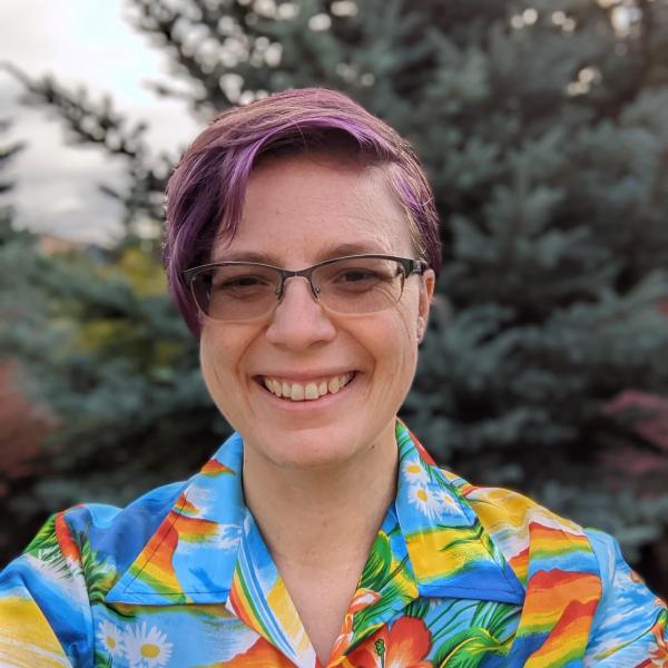 Woodros, wearing glasses and a bright, multi-colored shirt, is smiling at the camera, surrounded by green foliage in the background.