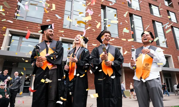 Four students wearing graduation regalia celebrate at Austin Hall among paper streamers