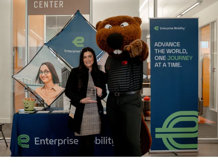 Ashley Parisi at COB's fall career fair. She stands in front of an Enterprise Mobility table with Benny Beaver. Both are wearing business casual apparel.